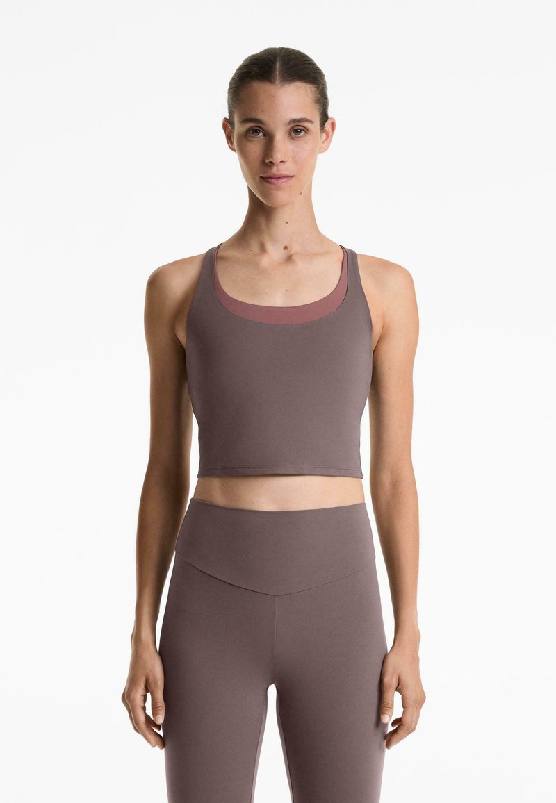 Kvinna som står framåtvänd, iklädd en mauve crop tank top och högmidjade leggings mot en vit bakgrund.