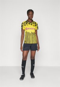 Žena stojaca v žltom a čiernom futbalovom drese Borussia Dortmund, čiernych šortoach, čiernych ponožkách po kolená a čiernych kopačkách na jednoduchom pozadí.