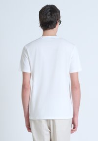 Camiseta de algodón blanca con mangas cortas, escote redondo y corte relajado. Presenta un sutil detalle de logo en la parte posterior del cuello.