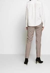Chemise blanche à manches longues avec une emmanchure dans le dos, associée à un pantalon à carreaux aux teintes rouge, noir et beige, et des bottines noires.
