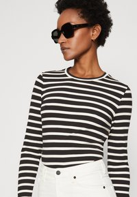 Maglietta a maniche lunghe a righe bianche e nere con texture a coste, abbinata a pantaloni bianchi. Occhiali da sole neri completano il look.