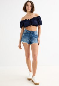 Top corto azul marino sin mangas con fruncido en la parte delantera y bordes con volantes, combinado con pantalones cortos de denim desgastados y deportivas blancas.