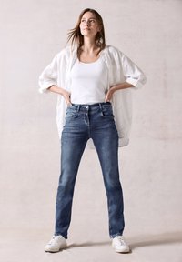 Vaqueros de mezclilla azul con un diseño ajustado, estilo de cinco bolsillos y cierre de botón. Se llevan con una camiseta sin mangas blanca y una camisa blanca de gran tamaño.