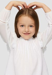 Blouse blanche à manches longues avec des détails froncés et de la broderie, au design plissé et avec un col rond. Jeune enfant posant les mains au-dessus de la tête.