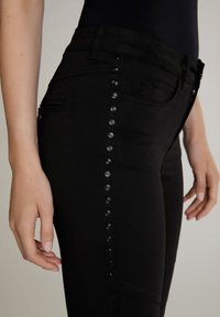 Zwarte jeans met een slim fit, zijzakken en een rij zwarte studs langs de buitennaad voor extra detail. Glad textuur en minimalistisch ontwerp.