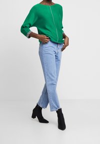 Groene losse trui met een bootneck en grijze boorden, gecombineerd met lichtblauwe rechte jeans en zwarte enkellaarsjes.