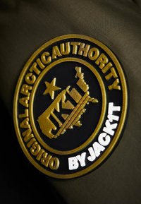 Okrągła czarno-złota naszywka z gwiazdą i napisem "Original Arctic Authority" oraz białym napisem "By Jacklt" na oliwkowej tkaninie.