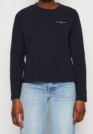 Person trägt ein marineblaues, langärmeliges Tommy Hilfiger Shirt mit Logo auf der Brust und hellblaue Jeans, steht vor einem schlichten Hintergrund.