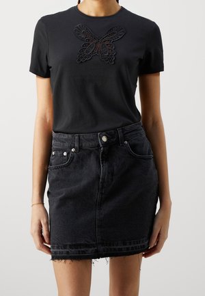 Kobieta ubrana w czarną koszulkę z krótkim rękawem z koronkowym motylem i czarną jeansową mini spódniczkę z wysokim stanem i postrzępionym brzegiem.