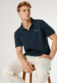 Mladý muž s červenými vlasmi sediaci na drevenej stoličke, oblečený v tmavomodrej polo košeli a svetlo béžových nohaviciach, hľadí na stranu.