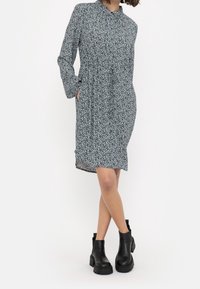 Blumenmusterkleid mit langen Ärmeln, hohem Ausschnitt und versteckten Seitentaschen. Dunkler Hintergrund mit einem hellblauen und weißen Blumenmuster.