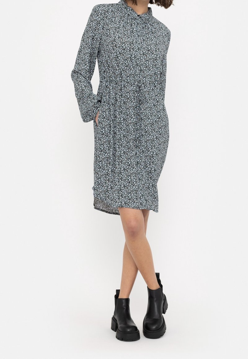 Blumenmusterkleid mit langen Ärmeln, hohem Ausschnitt und versteckten Seitentaschen. Dunkler Hintergrund mit einem hellblauen und weißen Blumenmuster.