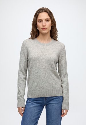 Jeune femme aux cheveux bruns ondulés portant un pull gris clair à manches longues et un jean bleu, debout devant un fond blanc uni.