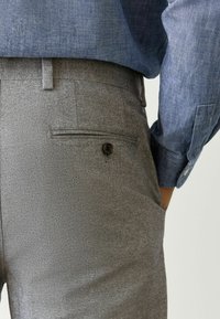 Grau gestreifte Hose mit maßgeschneidertem Schnitt, ausgestattet mit einer Gesäßtasche mit Knopfverschluss. Kombiniert mit einem blauen Hemd mit Knopfleiste.