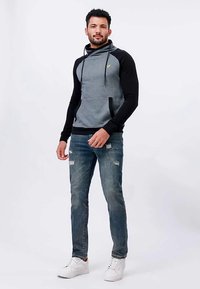Homem em pé, vestindo um hoodie preto e cinza, jeans azuis rasgados e sapatilhas brancas, com fundo branco simples.
