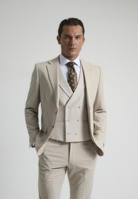 Traje beige de tres piezas con corte ajustado, chaleco de lino, camisa blanca y corbata marrón con un diseño estampado. Textura suave, diseño a medida.
