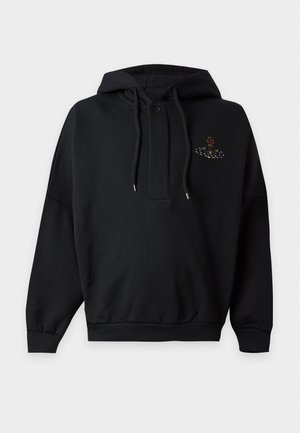 Hombre con sudadera negra con capucha y pequeño logo, pantalones negros sueltos con rayas verticales rojas y zapatos negros, de pie con una mano en el bolsillo.