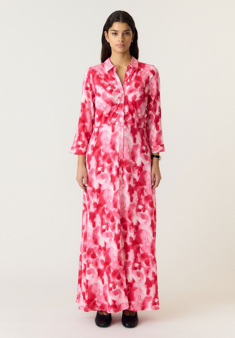 Vrouw staat gekleed in een lange roze en rode waterverfpatroon blousejurk met lange mouwen en zwarte schoenen op een lichte achtergrond.