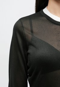 Mustast poolläbipaistvast materjalist pikkade varrukatega top, millel on valge soonikuga kaelus ja nurkadega disainielemendid kangal.