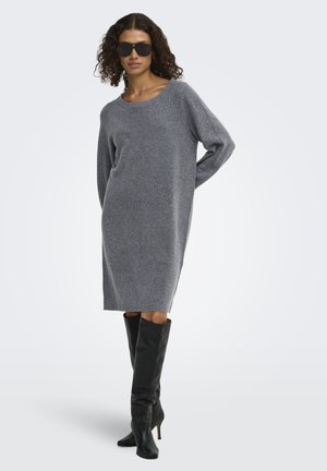 Femme aux cheveux bouclés portant une robe en maille grise à manches longues, des bottes noires jusqu'au genou et de grandes lunettes de soleil noires, debout devant un fond uni.