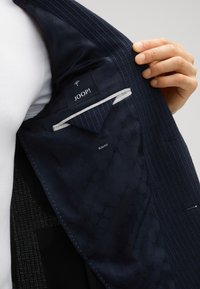Binnenkant van een marineblazer met textuurstof, inclusief een zak, een label met de tekst "JOOP!" en een "SLIM FIT" tag. Met een subtiel patroon.