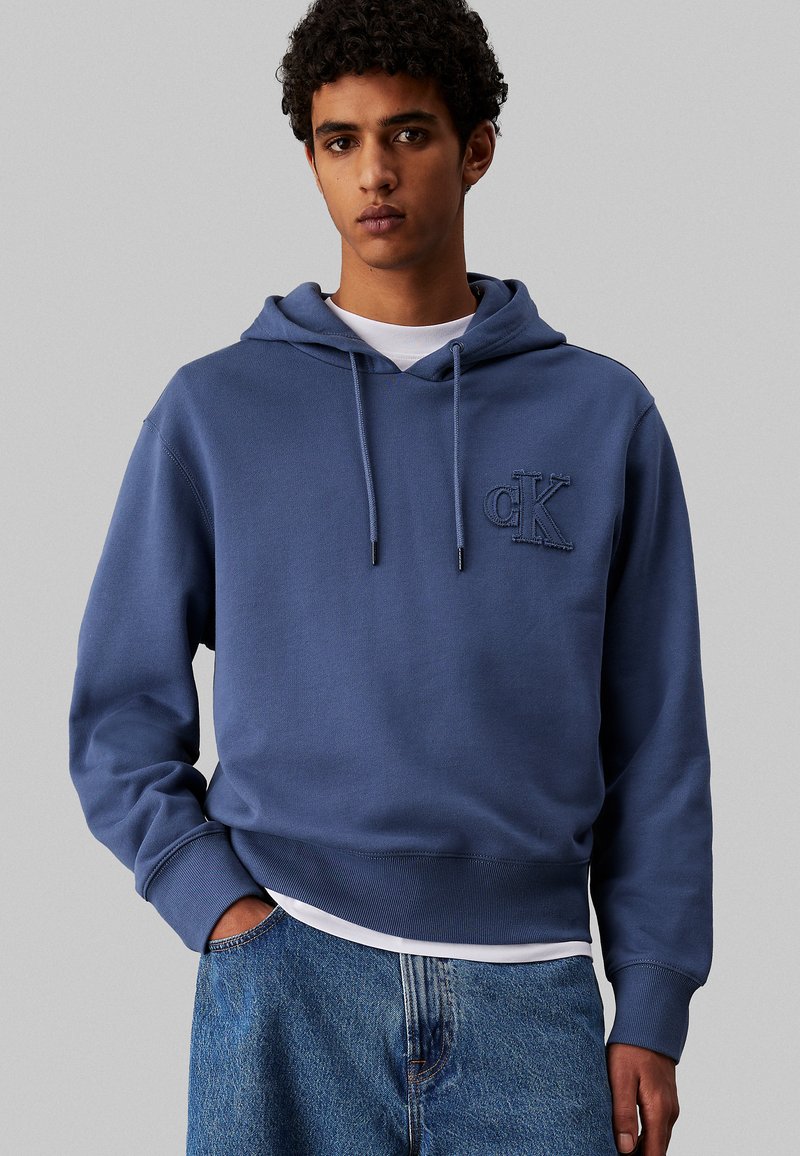 Felpa in cotone blu navy con cappuccio e coulisse, polsini e orlo a coste. Presenta un piccolo logo tono su tono sul lato sinistro del petto. Indossata sopra una camicia bianca.