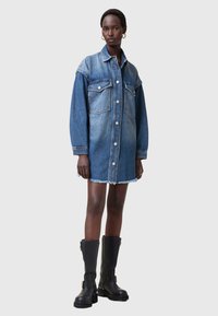 Denim krekli kleita, gaisā līdz tumši zilā gradientā, pogāta priekšpuse, divas krūšu kabatas, saplēsta apakša, valkāta ar melniem līdz ceļiem augstiem zābakiem.