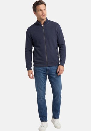 Man in een navy zip-up jack, blauwe jeans en witte sneakers, staand en iets naar rechts kijkend tegen een effen lichte achtergrond.