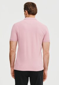 Różowa koszulka polo z krótkim rękawem o gładkiej fakturze. Posiada klasyczny kołnierzyk i prosty dół, noszona z ciemnymi spodniami. Widok z tyłu.