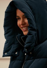 Chaqueta acolchada azul marino con una gran capucha. Presenta un cierre metálico y botones a presión. Textura suave con diseño acolchado, que proporciona calor.
