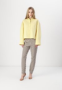 Cămașă galbenă scurtă, cu butoni, purtată împreună cu pantaloni slim-fit crem cu model. Persoană îmbrăcată cu tocuri bej, pe un fundal neted.