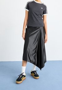 Žena v tmavě šedém tričku Adidas, černé asymetrické midi sukni, bílých ponožkách a černých teniskách Adidas se žlutými pruhy na modré podlaze.