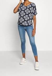 Granatowa bluzka z krótkim rękawem z białym wzorem kwiatowym, z dekoltem w serek. Jasnoniebieskie rurki i białe trampki dopełniają strój.