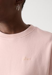 Lys rosa genser med ribbet krage og brodert logo i en lysere nyanse av rosa. Mykt stoff med en glatt tekstur.