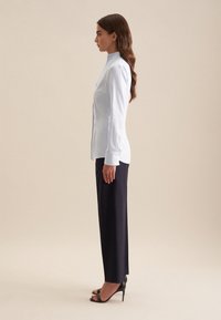 Camicia azzurra aderente con colletto alto, abbinata a pantaloni su misura blu scuro. Il modello indossa tacchi neri a strappo. Sfondo semplice.