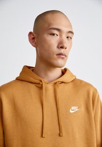 Brūns kapuces džemperis ar priekšējo kangaroo kabatu, savelcējiem un balto Nike logo kreisajā krūtī. Maiga auduma tekstūra.