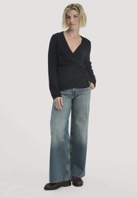 Frau mit kurzem blonden Haar, die ein schwarzes langärmeliges Wickeloberteil, weit geschnittene ausgewaschene blaue Jeans und dunkelbraune Slipper trägt, steht vor einem weißen Hintergrund.