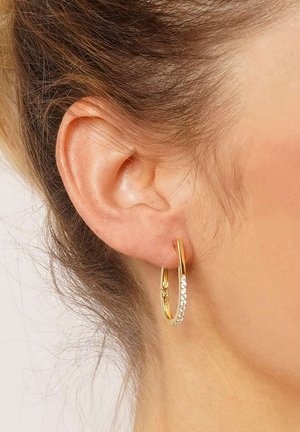 Goldene Creolen mit strukturiertem Finish und einer Reihe kleiner klarer Strasssteine auf einer Seite, die an einem durchstochenen Ohr befestigt sind.