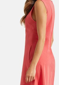 Robe sans manches longueur genoux en tissu corail, présentant une coupe droite et des poches latérales. Texture douce avec un léger éclat. Fermeture zippée au dos.