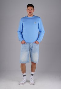 Helles blaues Sweatshirt mit Rundhalsausschnitt und kleinem Logo, kombiniert mit hellen Jeansshorts, weißen Turnschuhen und gemusterten Socken, vor einem grauen Hintergrund stehend.