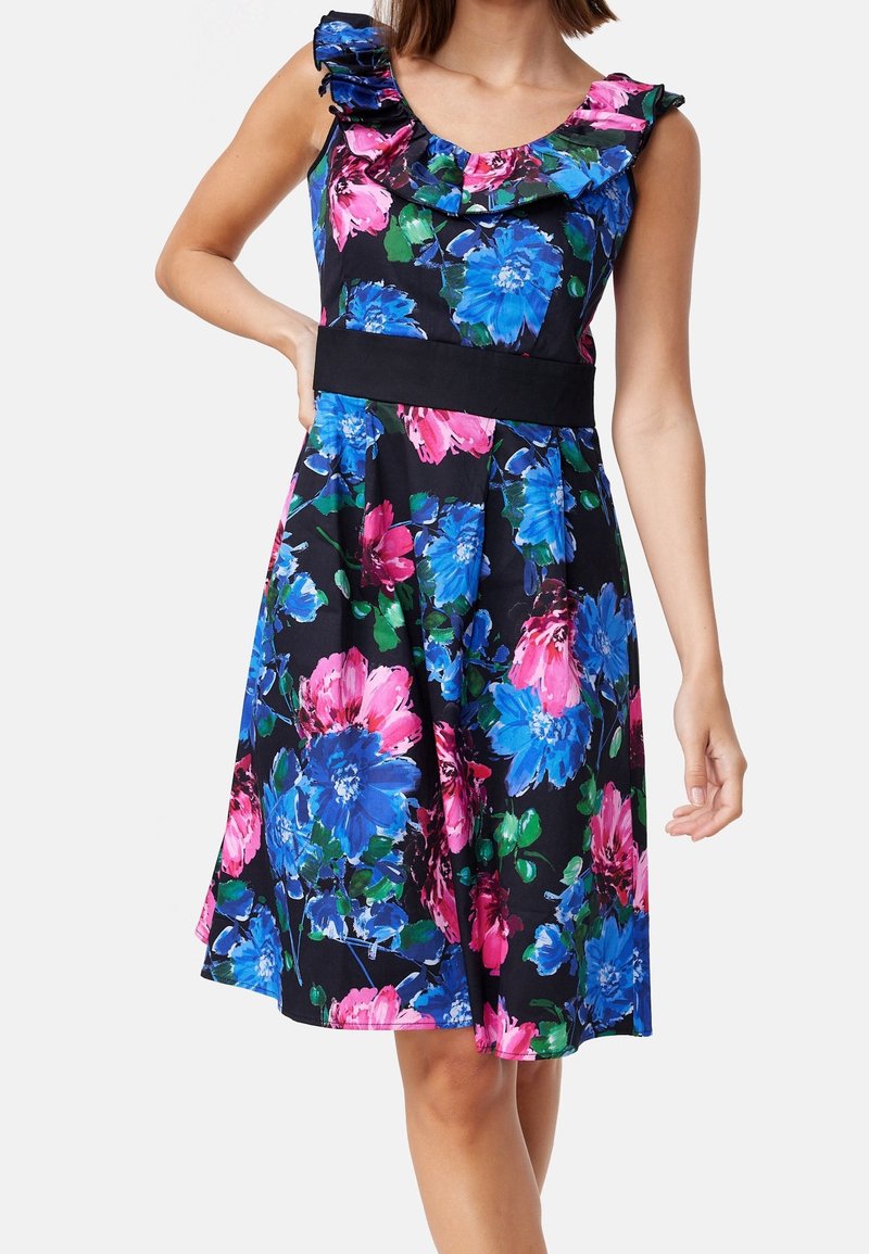 Robe sans manches mi-longue avec grand imprimé floral bleu et rose, encolure à volants, et large ceinture noire sur fond clair.