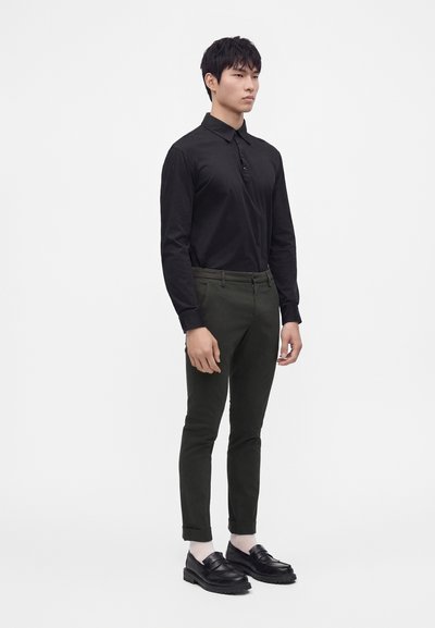 Homme portant une chemise noire à manches longues avec col, un pantalon slim vert foncé, des chaussettes blanches et des mocassins noirs, debout sur un fond blanc.