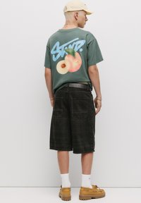 Grünes grafisches T-Shirt mit einem Pfirsichdesign und Oversized-Passform, kombiniert mit schwarzen knielangen Shorts und hellen Schuhen. Baumwollmischung.