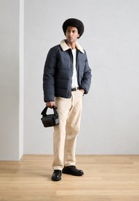 Puffjakke i marineblå med en kontrasterende cremefarvet krave, cremefarvede bukser og sorte sko. Holder en lille sort taske. Trægulv som baggrund.