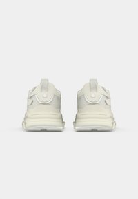 Par de zapatillas deportivas de color blanco roto vistas desde atrás, con acentos de malla, pestañas de tirón y suelas gruesas acolchadas sobre un fondo blanco liso.