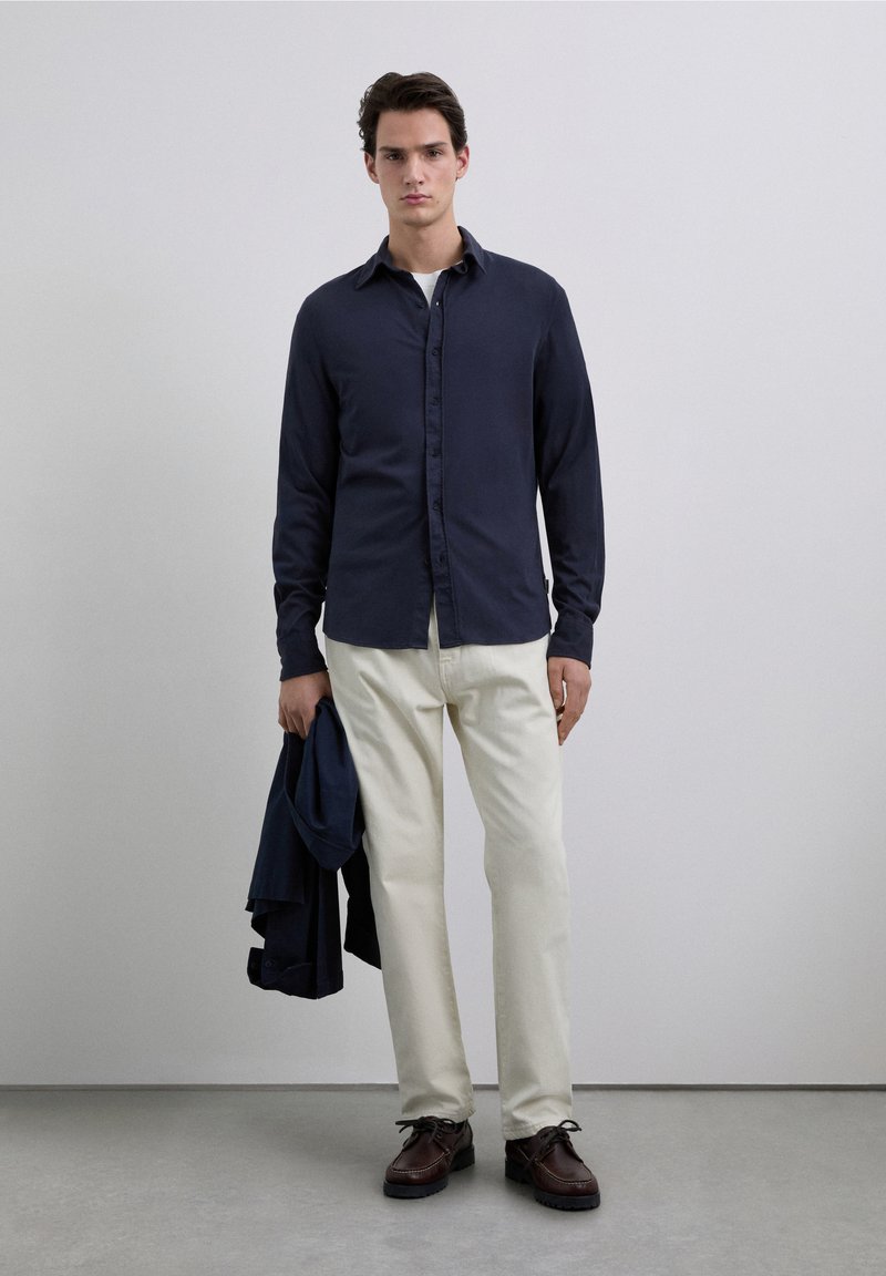 Navy long-sleeve button-up shirt, white undershirt, beige pants, holding a dark jacket, brown shoes with a thick sole, minimalist background.