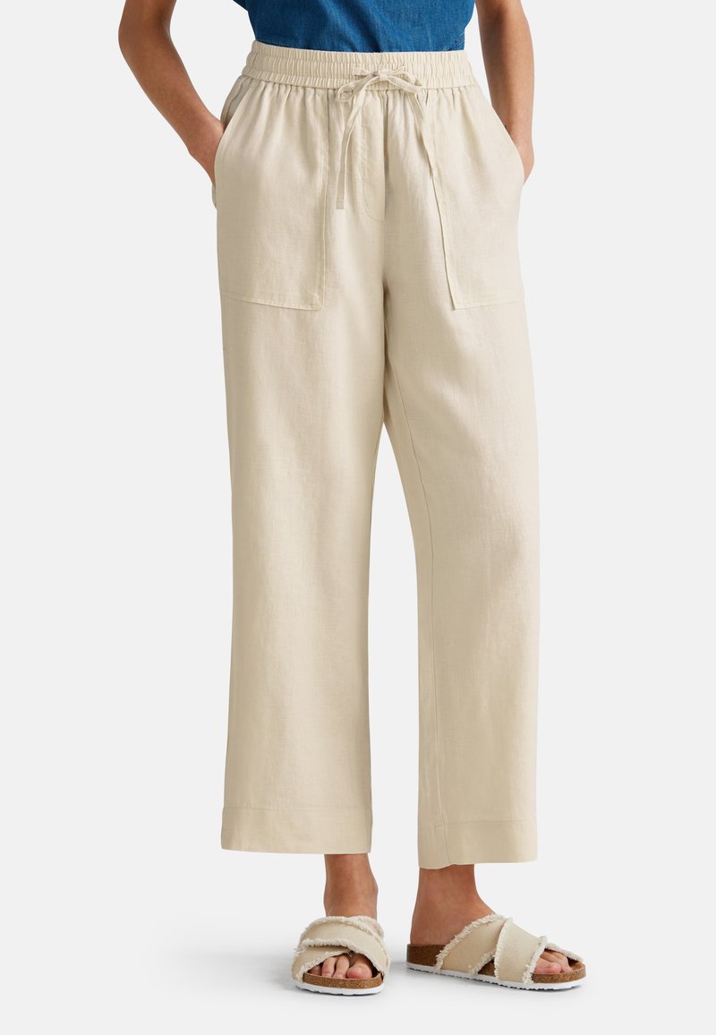 Beige Leinen-Hose mit weitem Bein, elastischem Bund, Kordelzug und zwei Fronttaschen. Getragen mit cremefarbenen Crossover-Sandalen.