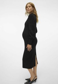Schwarzes Strickkleid mit strukturierten Puffärmeln und seitlichem Schlitz. Figurbetonte Silhouette, die für Komfort während der Schwangerschaft designed ist. Kombiniert mit spitzen Schuhen.