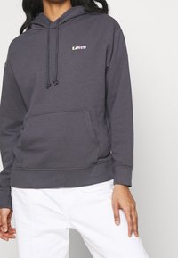 Personne portant un sweat à capuche Levi's gris foncé avec poche avant et cordons, associé à un pantalon blanc, debout devant un fond uni.
