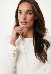 Bílý blazer s třemi texturovanými kulatými knoflíky na rukávu, spárovaný se světlým topem. Modelka má vlnité hnědé vlasy a neutrální líčení.